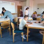 Schülerinnen und Schüler sitzen in kleinen Gruppen an Tischen und arbeiten konzentriert an einer Aufgabe. Im Hintergrund ist eine Projektion mit der Anleitung für das 'Spaghetti-Spiel' zu sehen. Die Aufgabe besteht darin, aus Spaghetti, Schnur und Klebeband einen Turm zu bauen, der einen Marshmallow trägt.