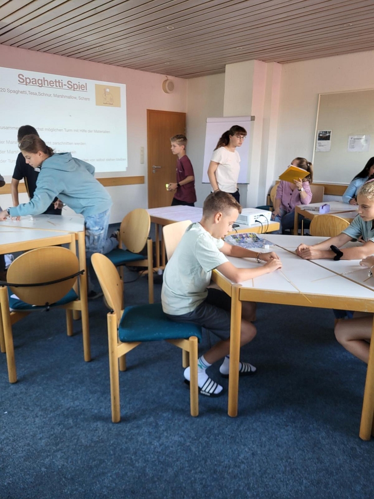 Schülerinnen und Schüler sitzen in kleinen Gruppen an Tischen und arbeiten konzentriert an einer Aufgabe. Im Hintergrund ist eine Projektion mit der Anleitung für das 'Spaghetti-Spiel' zu sehen. Die Aufgabe besteht darin, aus Spaghetti, Schnur und Klebeband einen Turm zu bauen, der einen Marshmallow trägt.