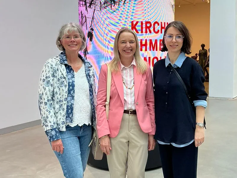 Drei lächelnde Frauen, Kolleginnen der Fachschaft Kunst, stehen nebeneinander in einer Galerie. Im Hintergrund ist eine große, leuchtende digitale Kunstinstallation mit pink-schwarzen Grafiken und Text zu sehen.