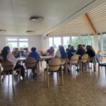 Das SV-Team sitzt in einer großen Runde an zusammengeschobenen Tischen in einem hellen Raum mit bodentiefen Fenstern. Die Gruppe, bestehend aus älteren Schülerinnen und Schülern, diskutiert und plant Aktionen. Auf den Tischen liegen Laptops, Tablets und Getränkeflaschen, während alle aufmerksam zuhören.