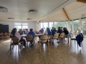 Das SV-Team sitzt in einer großen Runde an zusammengeschobenen Tischen in einem hellen Raum mit bodentiefen Fenstern. Die Gruppe, bestehend aus älteren Schülerinnen und Schülern, diskutiert und plant Aktionen. Auf den Tischen liegen Laptops, Tablets und Getränkeflaschen, während alle aufmerksam zuhören.