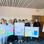 Eine Gruppe von neun jüngeren Schülerinnen und Schülern steht nebeneinander und hält stolz drei große, selbstgestaltete Plakate hoch. Die Plakate thematisieren Anti-Mobbing-Strategien. Darauf sind Slogans zu lesen wie 'Lacht mit, nicht aus!', 'Mobbing ist uncool' und 'Gemeinsam stark gegen Mobbing'.