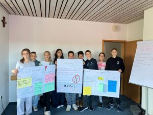 Eine Gruppe von neun jüngeren Schülerinnen und Schülern steht nebeneinander und hält stolz drei große, selbstgestaltete Plakate hoch. Die Plakate thematisieren Anti-Mobbing-Strategien. Darauf sind Slogans zu lesen wie 'Lacht mit, nicht aus!', 'Mobbing ist uncool' und 'Gemeinsam stark gegen Mobbing'.