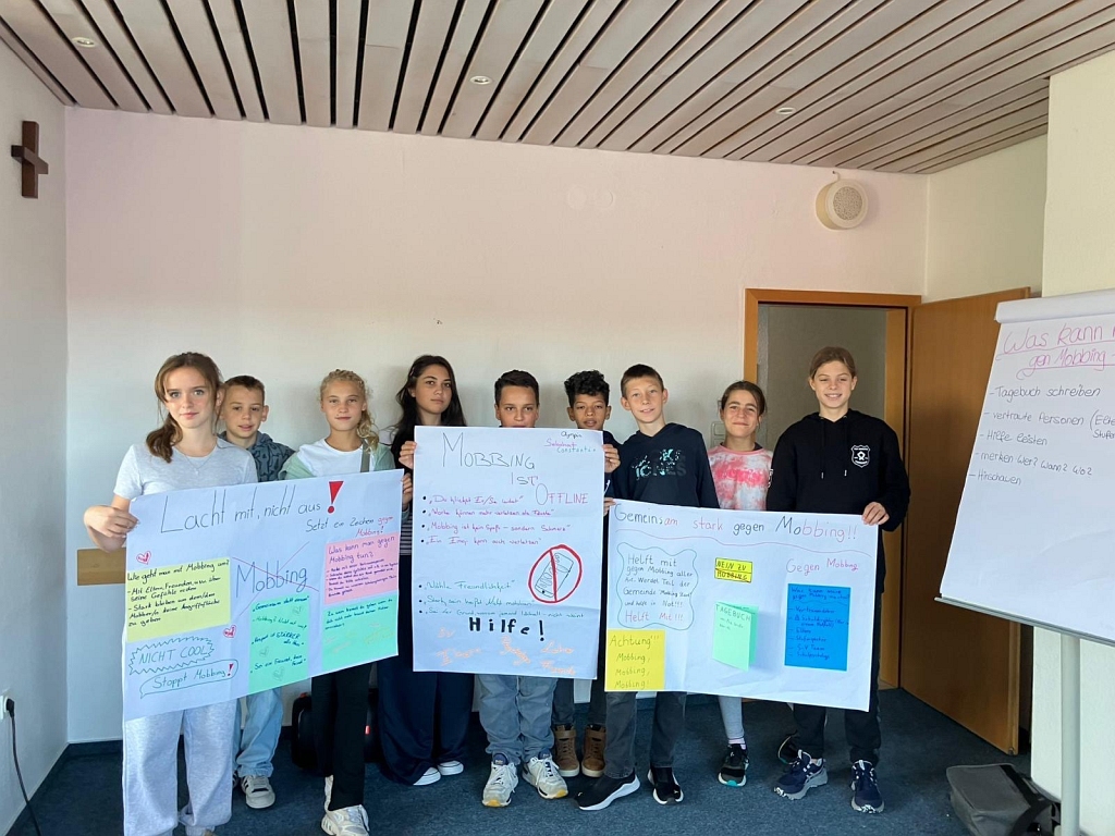 Eine Gruppe von neun jüngeren Schülerinnen und Schülern steht nebeneinander und hält stolz drei große, selbstgestaltete Plakate hoch. Die Plakate thematisieren Anti-Mobbing-Strategien. Darauf sind Slogans zu lesen wie 'Lacht mit, nicht aus!', 'Mobbing ist uncool' und 'Gemeinsam stark gegen Mobbing'.