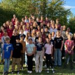 Ein Gruppenbild des SV-Teams 2025 im Freien. Etwa 50 Schülerinnen und Schüler verschiedener Altersstufen stehen gestaffelt auf einer Wiese vor einer Baumreihe. Viele lächeln in die Kamera und einige tragen dunkelrote T-Shirts. Das Wetter ist sonnig mit blauem Himmel.