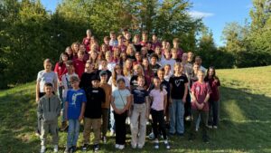 Ein Gruppenbild des SV-Teams 2025 im Freien. Etwa 50 Schülerinnen und Schüler verschiedener Altersstufen stehen gestaffelt auf einer Wiese vor einer Baumreihe. Viele lächeln in die Kamera und einige tragen dunkelrote T-Shirts. Das Wetter ist sonnig mit blauem Himmel.