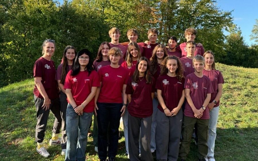 Gruppenfoto der gewählten Schul- und Stufensprecherinnen und -sprecher zusammen mit der Verbindungslehrerin Frau Wörner. Die Gruppe von etwa 17 Personen steht draußen auf einer Wiese vor grünen Bäumen und blauem Himmel. Fast alle tragen einheitliche dunkelrote T-Shirts mit einem weißen Logo auf der Brust, um ihren Zusammenhalt als Team zu zeigen.