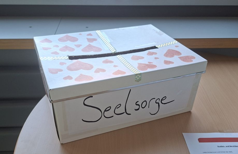 Eine weissfarbige Box mit pinkfarbenem Herzen bedruckt, darauf steht der Text "Seelsorge" in schwarzen Großbuchstaben.